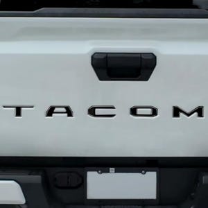 May include: The white tailgate of a pickup truck features the black lettering TACOM centred horizontally. A black handle is positioned above the text, and a rectangular number plate frame is located at the bottom centre of the vehicle.