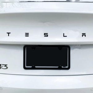 May include: A white car's rear with the black letters spelling "TESLA" across the top. A black license plate frame is centered below the letters. The car model "3" is visible on the left side.