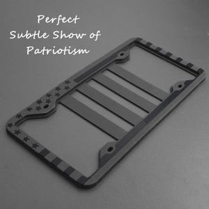 May include: A black license plate frame with a subtle American flag design. The frame has a raised edge and a smooth finish.