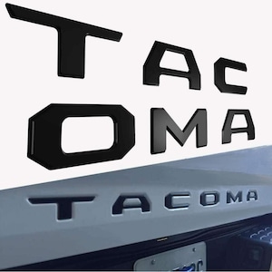 May include: Black plastic letters that spell out "TACOMA" in all caps. The letters are shown on a white surface and on the back of a silver truck.