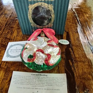 May include: A Waterford Holiday Heirloom ornament featuring a red, white, and green glass dove design. The ornament is in a box with the Waterford logo. The text on the box reads "Waterford Holiday Heirlooms". The text on the card reads "Care and Handling".