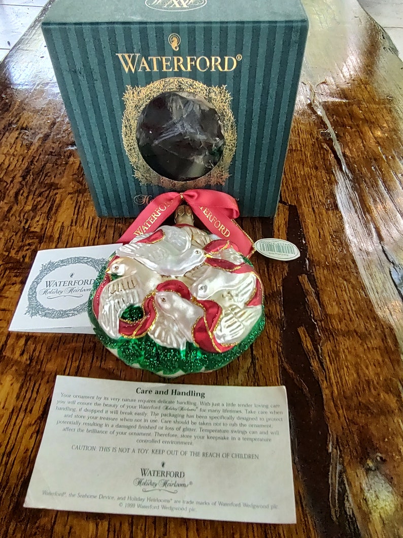 May include: A Waterford Holiday Heirloom ornament featuring a red, white, and green glass dove design. The ornament is in a box with the Waterford logo. The text on the box reads "Waterford Holiday Heirlooms". The text on the card reads "Care and Handling".