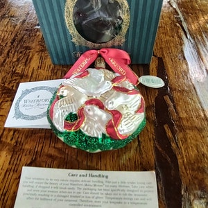 May include: A Waterford Holiday Heirloom ornament featuring a white glass dove with red and green accents. The ornament is surrounded by a green wreath and has a red ribbon.