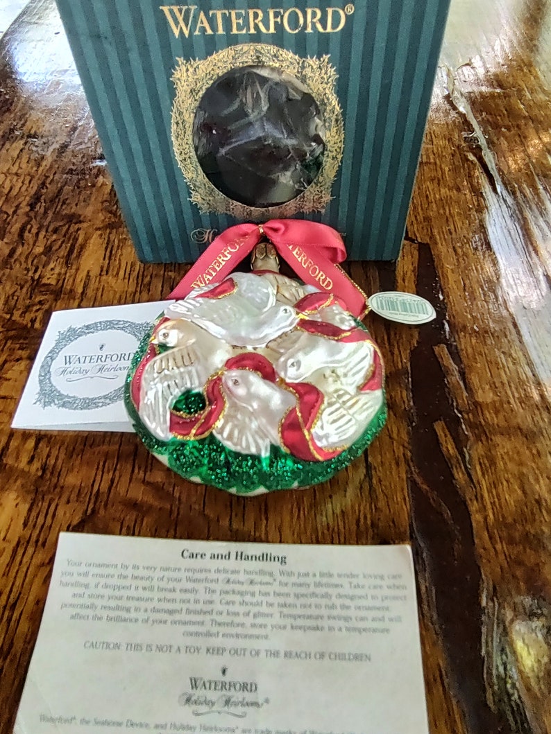 May include: A Waterford Holiday Heirloom ornament featuring a white glass dove with red and green accents. The ornament is surrounded by a green wreath and has a red ribbon.