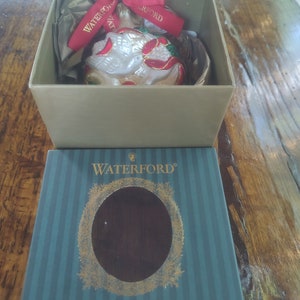 May include: A Waterford Holiday Heirlooms ornament box with a green and white striped design. The box features a gold wreath with a clear window showing a white glass ornament with a dove design. The ornament is decorated with red and gold accents.