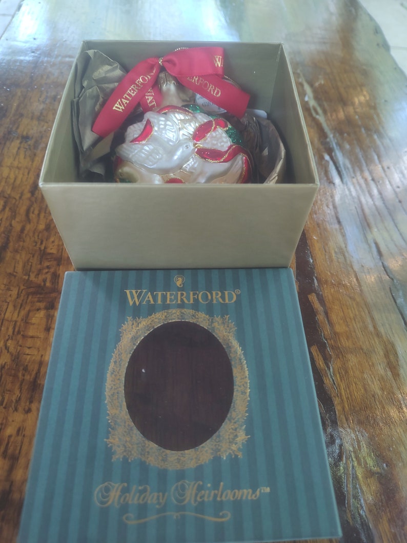 May include: A Waterford Holiday Heirlooms ornament box with a green and white striped design. The box features a gold wreath with a clear window showing a white glass ornament with a dove design. The ornament is decorated with red and gold accents.
