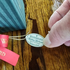May include: A green and white label with the text "Waterford Holiday Heirlooms Handmade in Poland" attached to a red ribbon.