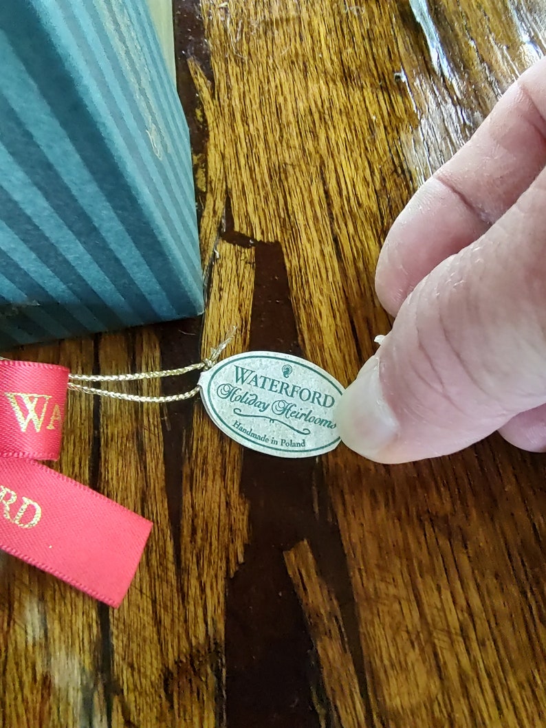 May include: A green and white label with the text "Waterford Holiday Heirlooms Handmade in Poland" attached to a red ribbon.