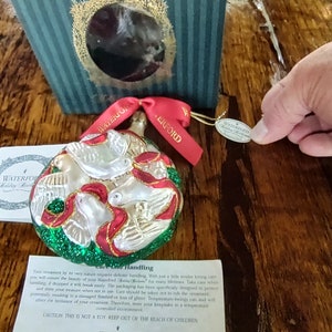 May include: A Waterford Holiday Heirlooms ornament featuring three white doves with red and gold accents on a green background. The ornament is in its original box with a tag that reads "Waterford Holiday Heirlooms".