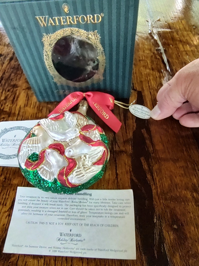 May include: A Waterford Holiday Heirlooms ornament featuring three white doves with red and gold accents on a green background. The ornament is in its original box with a tag that reads "Waterford Holiday Heirlooms".