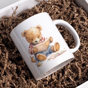 May include: White ceramic mug with a brown teddy bear reading a book. The bear is wearing a red and white striped shirt and blue jeans. The mug has a handle on the right side.