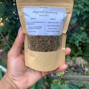 May include: A brown paper pouch with a white label that reads "Thyroid Soothing Herbal Tea Blend". The label lists the ingredients: Organic Lemon Balm, Organic Stinging Nettle, Organic Turmeric, and Organic Hibiscus Flowers. The pouch contains a blend of dried herbs.