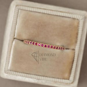 Könnte beinhalten: Ein dünner Ring aus Roségold mit einer durchgehenden Reihe kleiner, runder, leuchtend pinker Edelsteine auf der Oberseite. Der Ring befindet sich in einer quadratischen, beigen Samtschmuckschatulle mit dem aufgedruckten Diamond Vibe Logo.