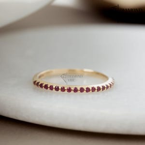 Puede incluir: Un anillo de banda fina de oro con una hilera continua de pequeños rubíes redondos de color rojo intenso engastados en la parte frontal. El metal tiene un acabado pulido y el texto DIAMOND VIBE es visible en el centro del anillo.