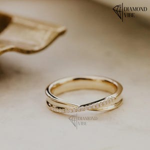May include: A gold ring featuring a crossover design where one band is plain polished gold and the other is encrusted with a row of small clear stones. The ring is marked with the text DIAMOND VIBE in the upper right and lower center.