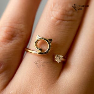 May include: A gold-colored ring featuring a cat-shaped design with a heart-shaped gemstone. The ring wraps around the finger, with the cat's head forming a loop. The gemstone is set on the opposite side of the ring.