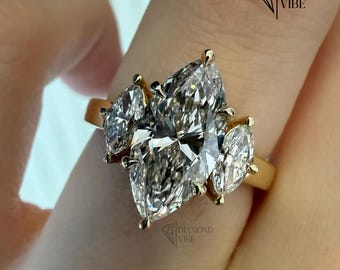 May include: A gold ring featuring a large central marquise-cut diamond flanked by two smaller marquise-cut diamonds on each side. The setting is polished yellow gold, and the stones are clear and faceted. The text DIAMOND VIBE is visible.