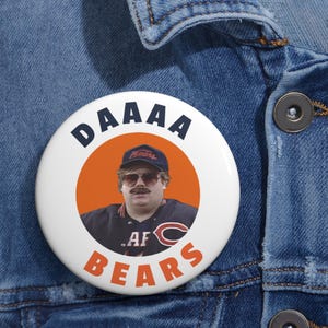 Op de afbeelding: Een witte knop met een afbeelding van een man die een Chicago Bears-pet en -shirt draagt. De knop zegt "DAAAA BEARS" in een cirkel rond de afbeelding.