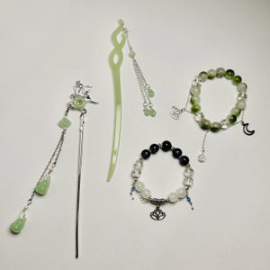 Puede incluir: Un conjunto de tres pulseras de cuentas verdes y blancas con dijes de plata. Las pulseras presentan una luna creciente, una flor de loto y una pequeña flor. Las pulseras están hechas con diferentes tipos de cuentas, incluyendo cuentas transparentes, verdes y blancas.