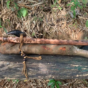 May include: A carved wooden flute with a dark bird-shaped head, lying on a wooden log. The flute features detailed patterns and a beaded cord. The natural wood grain is visible. The flute's design suggests it is a traditional musical instrument.