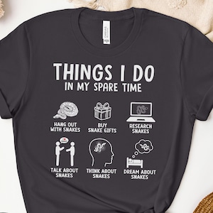 Op de afbeelding: Donkergrijs T-shirt met de tekst "Things I do in my spare time" en een lijst met activiteiten die te maken hebben met slangen. De activiteiten omvatten: tijd doorbrengen met slangen, slangencadeaus kopen, onderzoek doen naar slangen, praten over slangen, denken aan slangen en dromen over slangen.