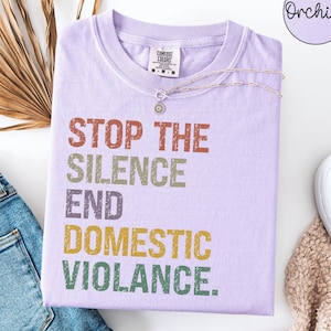 Può includere: Una t-shirt viola chiaro con il testo usurato e multicolore STOP THE SILENCE END DOMESTIC VIOLENCE. Il capo presenta un girocollo e un'etichetta del marchio Comfort Colors sul colletto interno. Una catenina d'argento poggia sul tessuto.