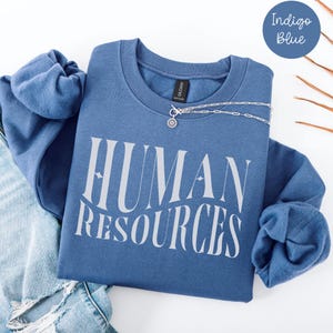 Peut inclure: Sweat-shirt bleu indigo avec l'inscription "HUMAN RESOURCES" en blanc dans une police stylisée. Un collier argenté avec un pendentif est posé sur le sweat-shirt. Le sweat-shirt est plié sur un jean bleu.