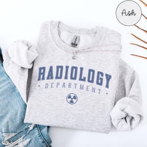 May include: A gray sweatshirt with the text "Radiology Department" in blue lettering and a blue radiation symbol. The sweatshirt is on a white background.