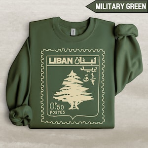 Op de afbeelding: Militairgroen sweatshirt met een beige stempelontwerp. De stempel toont het woord "LIBAN" in het Engels en Arabisch, een boomillustratie en de tekst "0.50 POSTES". De sweatshirt heeft een ronde hals en lange mouwen.