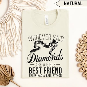 Op de afbeelding: Een lichtbeige T-shirt met een zwarte afbeelding waarop staat "Whoever said diamonds are a girls best friend never had a ball python". De afbeelding bevat ook een zwart-witte illustratie van een slang.