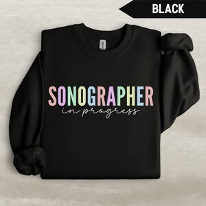 Może przedstawiać: Czarna bluza z napisem "SONOGRAPHER in progress" w kolorach tęczy.