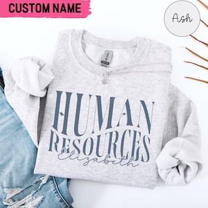 Könnte beinhalten: Ein grauer Pullover mit dem Aufdruck "HUMAN RESOURCES Elizabeth" in Blau auf der Vorderseite. Der Pullover ist gefaltet und auf einem blauen Jeansstoff-Hintergrund abgelegt.