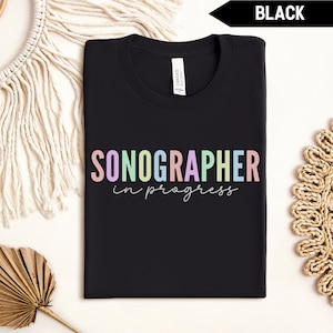 Puede incluir: Camiseta negra con el texto "SONOGRAPHER in progress" en colores arcoíris.