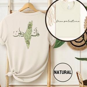 Puede incluir: Una camiseta beige con un contorno verde de Palestina y texto árabe que dice "Free Palestine". La parte posterior de la camiseta tiene el mismo diseño con la adición de ramas de olivo y flores moradas. La camiseta está en una percha y tiene una etiqueta "NATURAL".