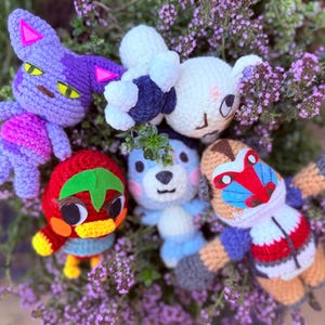 May include: A collection of handmade crochet plush toys in various colors. The toys include a purple cat, a white bear, a red bird, a blue bear, and a monkey. The toys are surrounded by purple flowers.