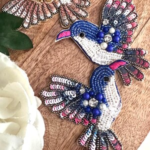May include: Three embroidered hummingbird patches featuring iridescent sequins and beads. The hummingbirds are in shades of pink, blue, white, and gold.