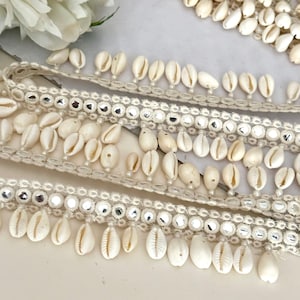 以下が含まれることがあります： 銀色のビーズレースのベースに白い貝殻のトリム。トリムは小さな白い貝殻のフリンジが付いています。