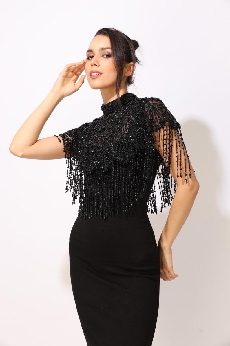Model wearing a black embroidered beaded cape with scalloped edges and fringe. The sheer net base is hand-embellished with intricate embroidery and sparkling black beads, creating a statement bridal or evening accessory.