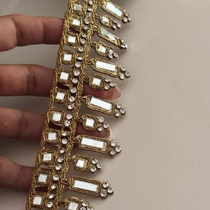 Puede incluir: Borde dorado con cuadrados espejados y pedrería transparente. El borde tiene una franja de rectángulos espejados.