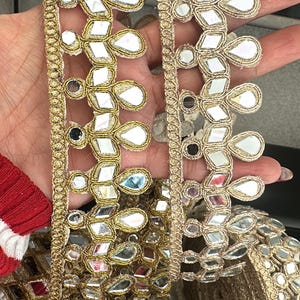 May include: Gold trim with mirror embellishments, rolled up and held in a hand. This decorative trim is perfect for adding a touch of sparkle to any project.