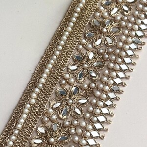 Gold embroidered lace with pearl and mirror detailing, laid flat on white surface with floral decor, for luxury craft designs.