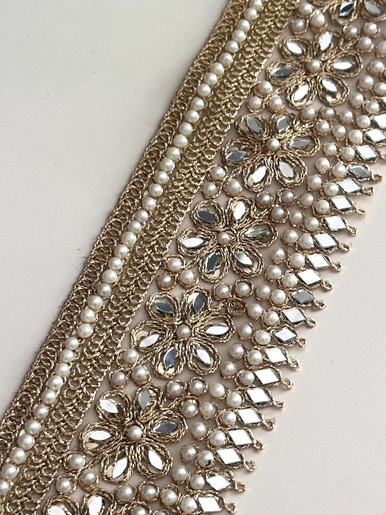 Gold embroidered lace with pearl and mirror detailing, laid flat on white surface with floral decor, for luxury craft designs.