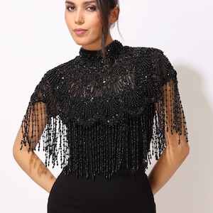 Model wearing a black embroidered beaded cape with scalloped edges and fringe. The sheer net base is hand-embellished with intricate embroidery and sparkling black beads, creating a statement bridal or evening accessory.