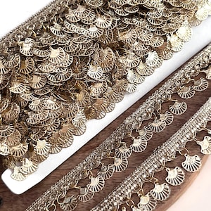May include: Close-up of gold-colored decorative trim with a fan-shaped design. The trim is layered, with a dense top layer and a dangling fringe. The trim is likely used for embellishing clothing or crafts.
