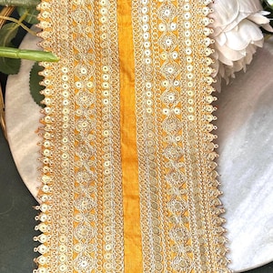 May include: A yellow fabric trim with a decorative design of silver sequins and embroidery. The trim is approximately 45 cm long and has a scalloped edge.