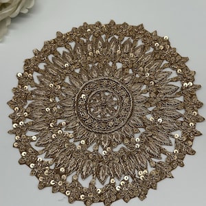 Peut inclure: Applique brodée circulaire de couleur or avec un motif floral. L'applique présente des détails complexes de feuilles et de paillettes, créant un embellissement décoratif et orné. Le motif est centré avec un motif en spirale.