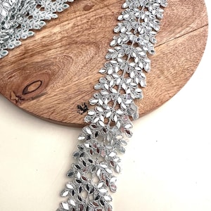 Puede incluir: Borde plateado con un diseño de hojas con lentejuelas de espejo. El borde está sobre una superficie de madera.