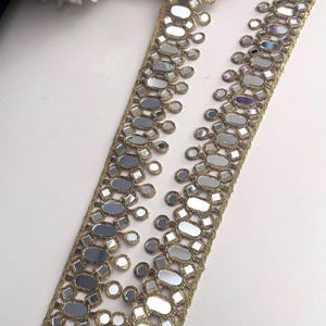 Può includere: Bordura dorata con inserti a specchio. La bordura presenta un motivo ripetuto di specchi ovali e quadrati, creando un elemento riflettente e decorativo. La bordura viene probabilmente utilizzata per abbellire abiti o progetti artigianali.