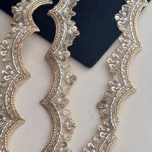 May include: Close-up of ornate trim with a scalloped edge. The trim features gold and silver metallic threads, small white beads, and sequin accents. The design is intricate and detailed, suitable for embellishing garments or crafts.