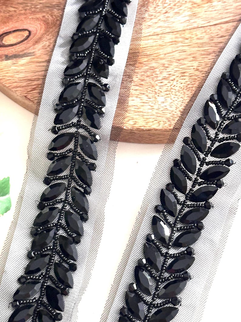 May include: Black beaded trim with leaf-shaped embellishments on a sheer white fabric. The trim is perfect for adding a touch of elegance to any project.